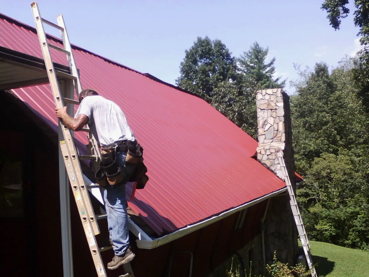 Roofer on a ladder beside a red metal roof and stone chimney — Soffit & Fascia Repair by Roofing Brothers in Karthaus